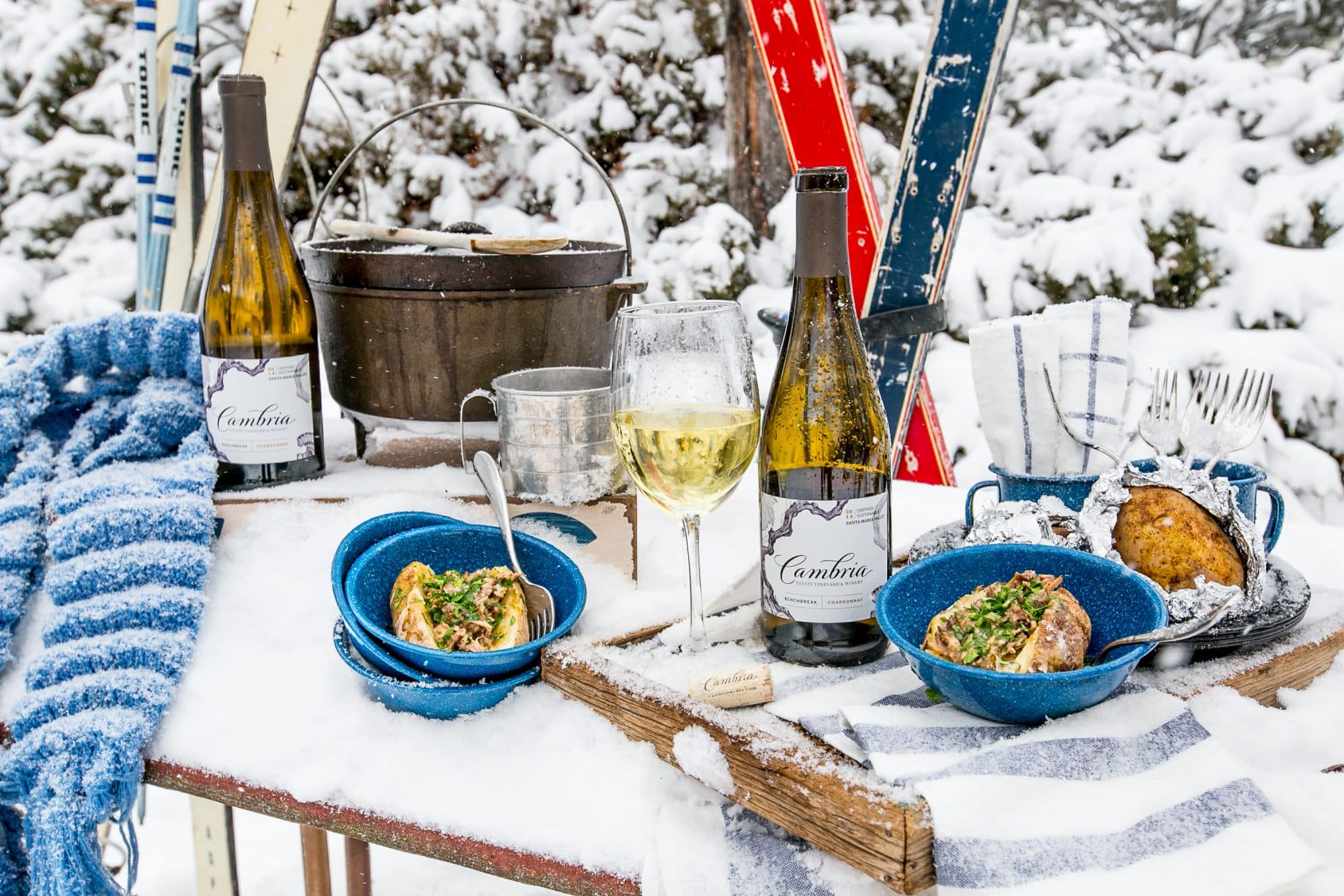 snow covered table with Cambria wine, country rib stuffed potatoes in bowls and skies in the background