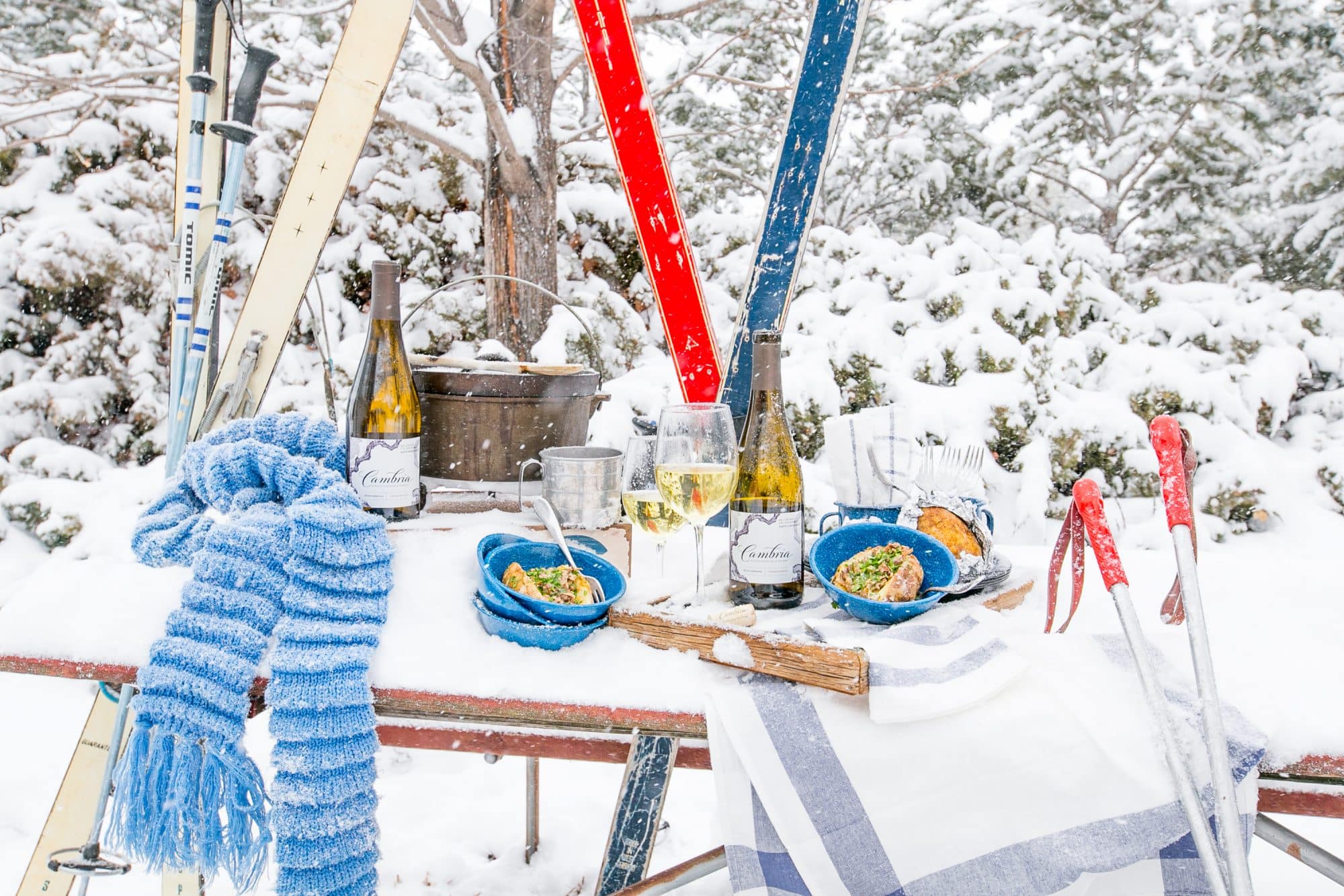 snow covered table with Cambria wine, country rib stuffed potatoes in bowls and skies in the background