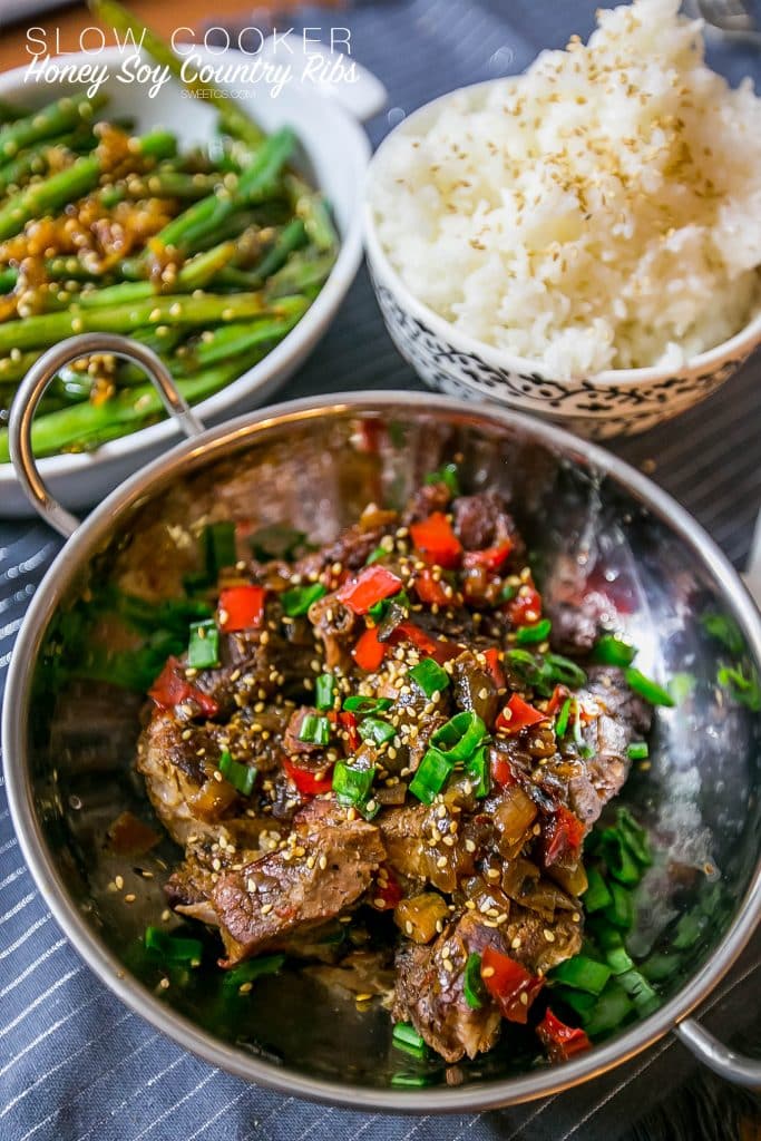 asian ribs with sesame seeds and green onions and veggies in a metal bowl, rice, and green beans in the background.
