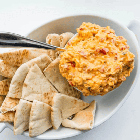 A white bowl containing pita bread slices and a serving of pimiento cheese dip, with a spoon resting inside.