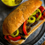A Vegan Philly Cheese Steak Hoagie on a crusty roll, filled with grilled mushrooms, red bell peppers, and sliced banana peppers, served on a dark plate.