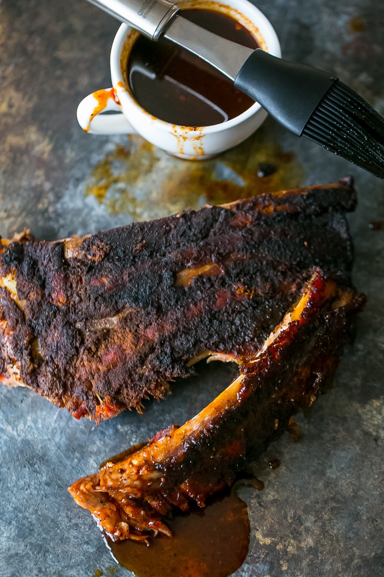 lovely fall off the bone ribs on a black surface with sauce in the background with sauce in a mug and a silicone brush.