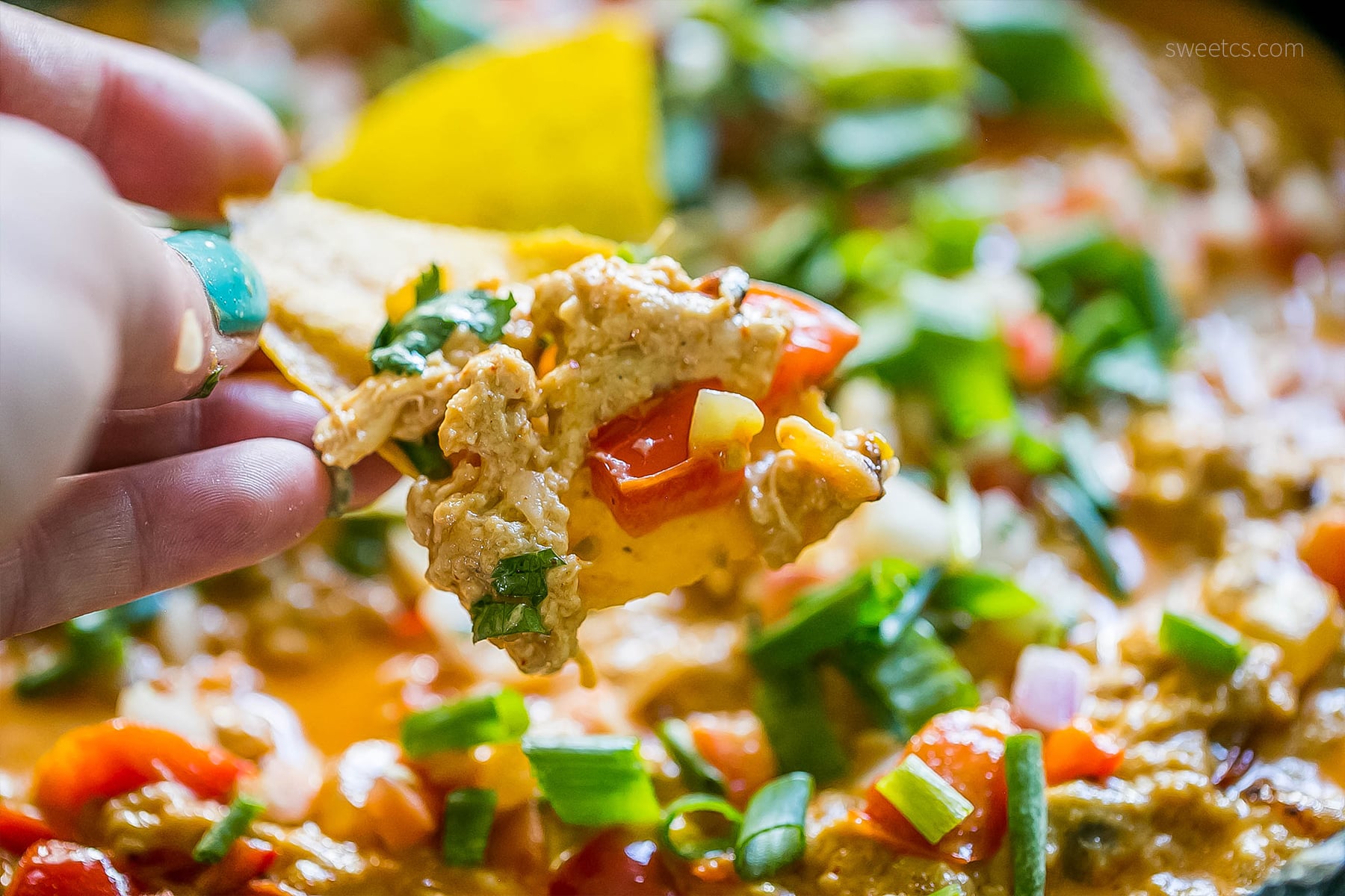 a chip going into dip with chicken, green onions, tomato, red onion, cheesy, and sauce in it