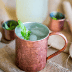 A copper mug containing a chilled Virgin Mule garnished with a basil leaf, placed on a rustic burlap surface with another mug and a pitcher in the background.