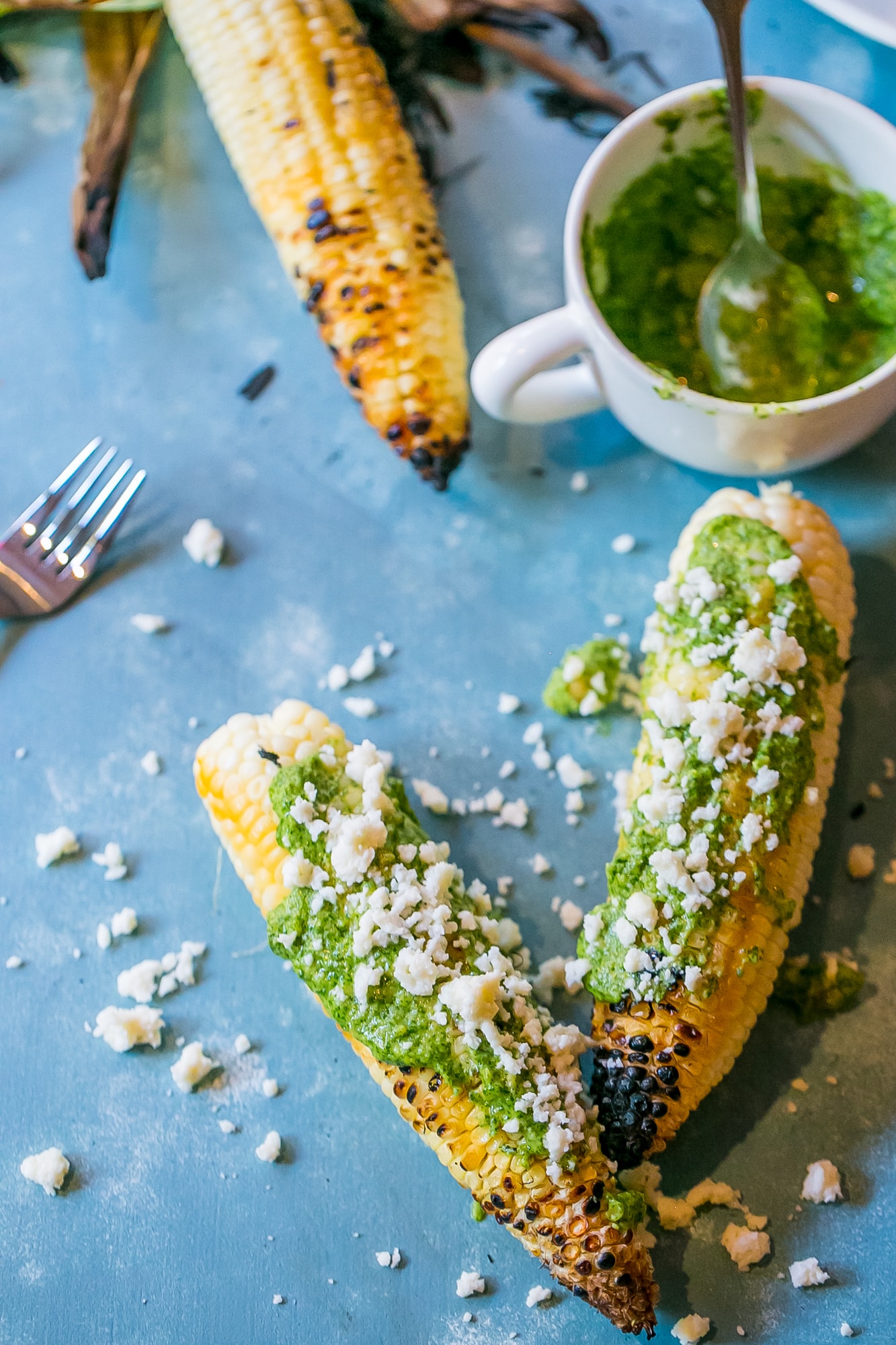 grilled corn with green pesto and crumbled cheese on it, mug of pesto in the background