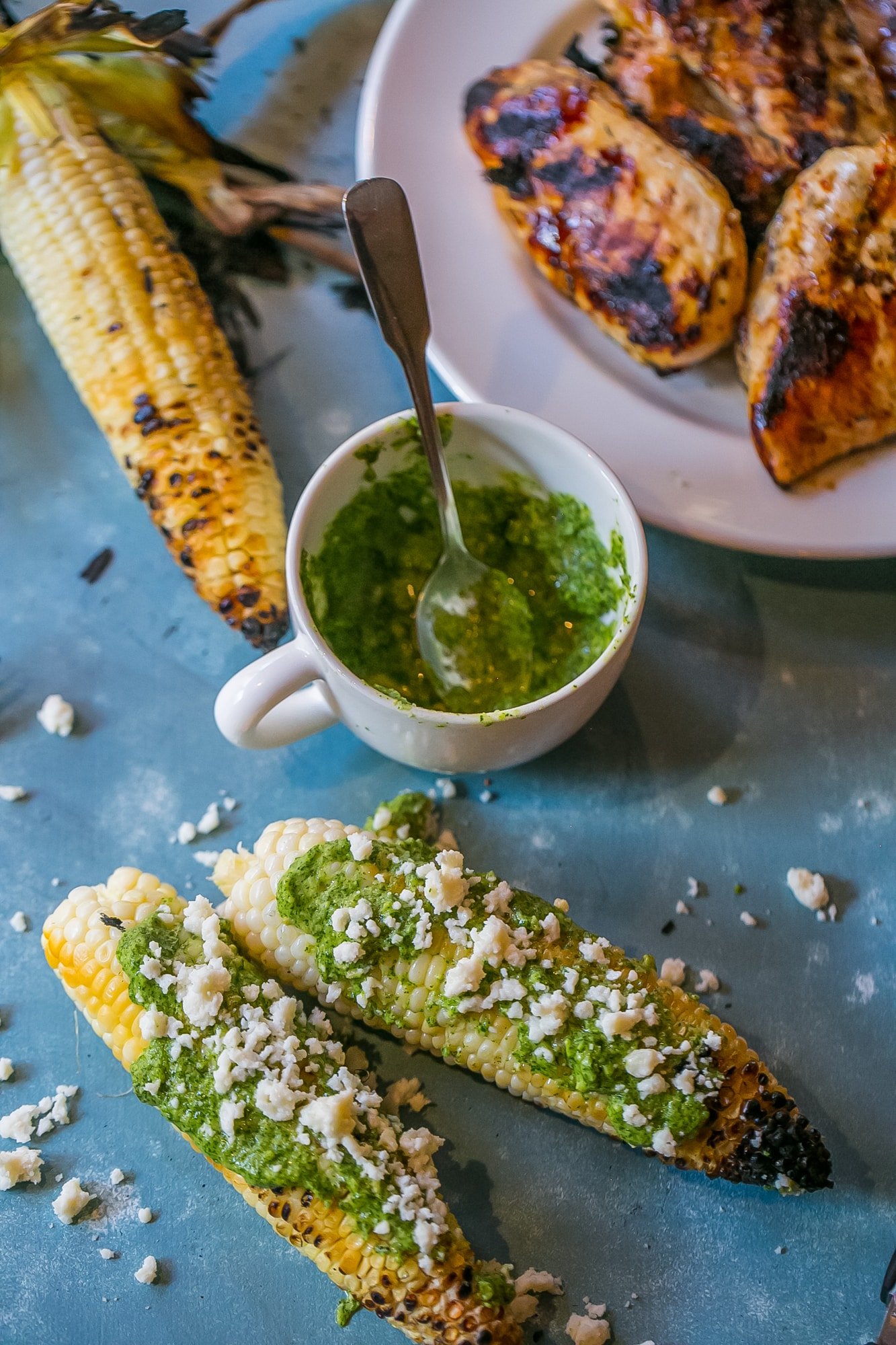 grilled corn with green pesto and crumbled cheese on it, mug of pesto in the background