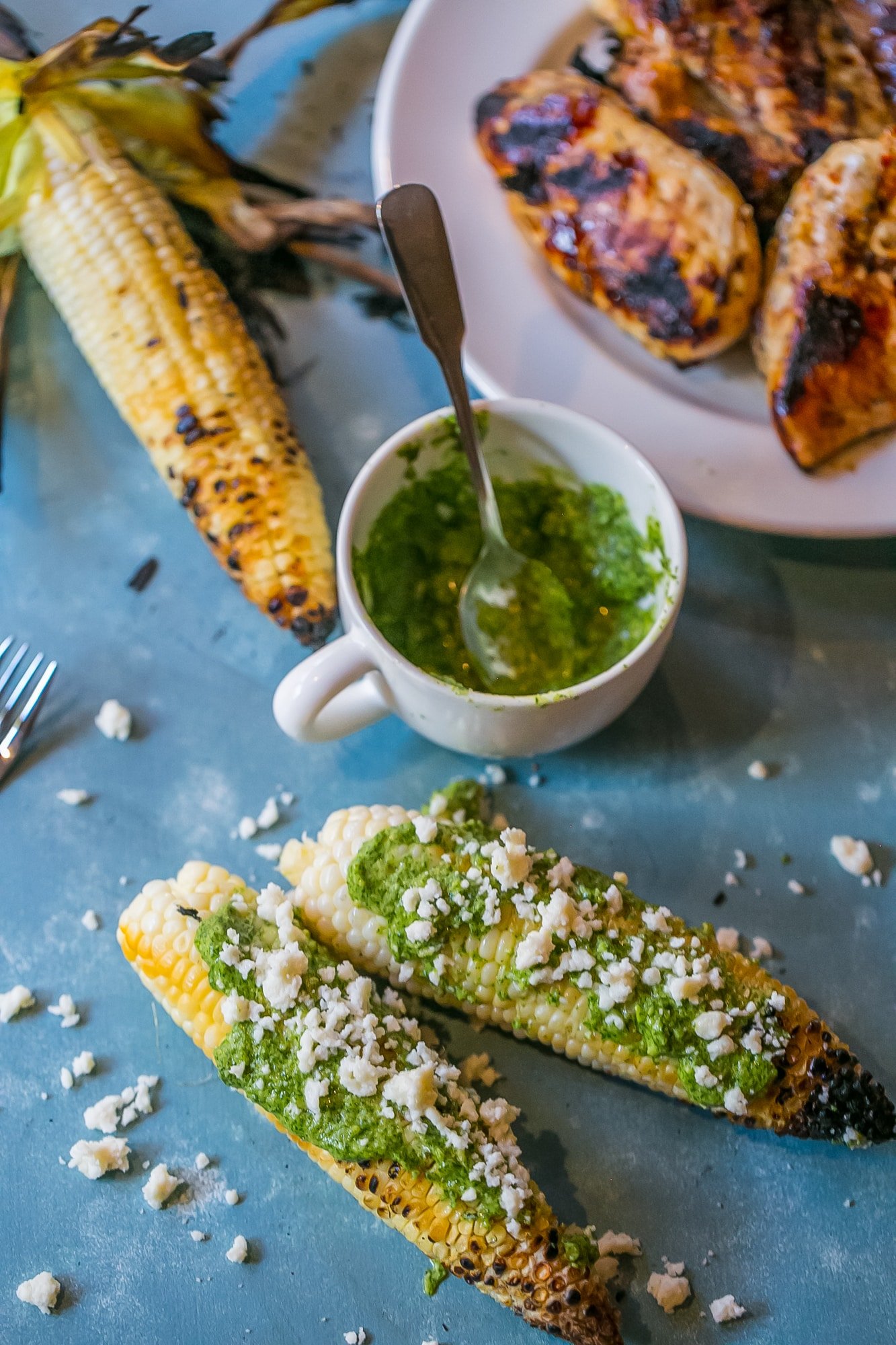 grilled corn with green pesto and crumbled cheese on it, mug of pesto in the background