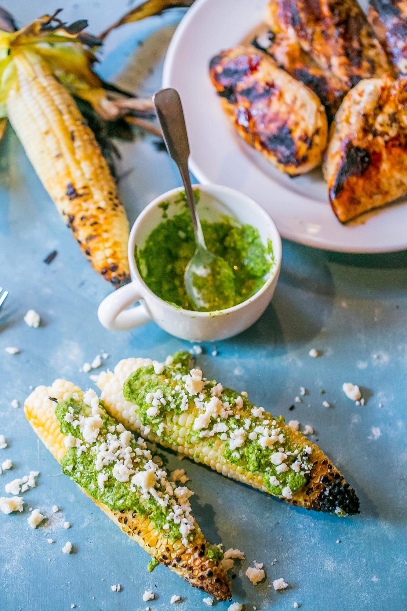 grilled corn with green pesto and crumbled cheese on it, mug of pesto in the background
