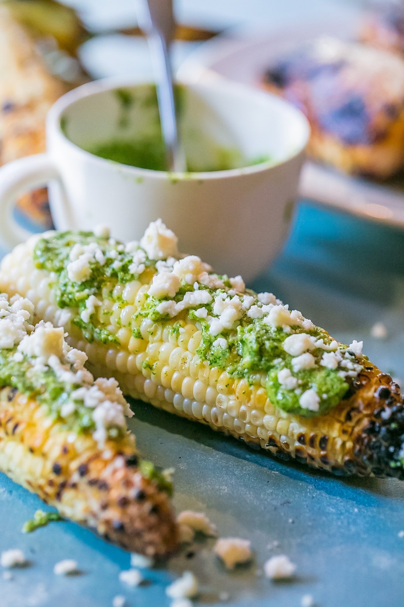 grilled corn with green pesto and crumbled cheese on it, mug of pesto in the background