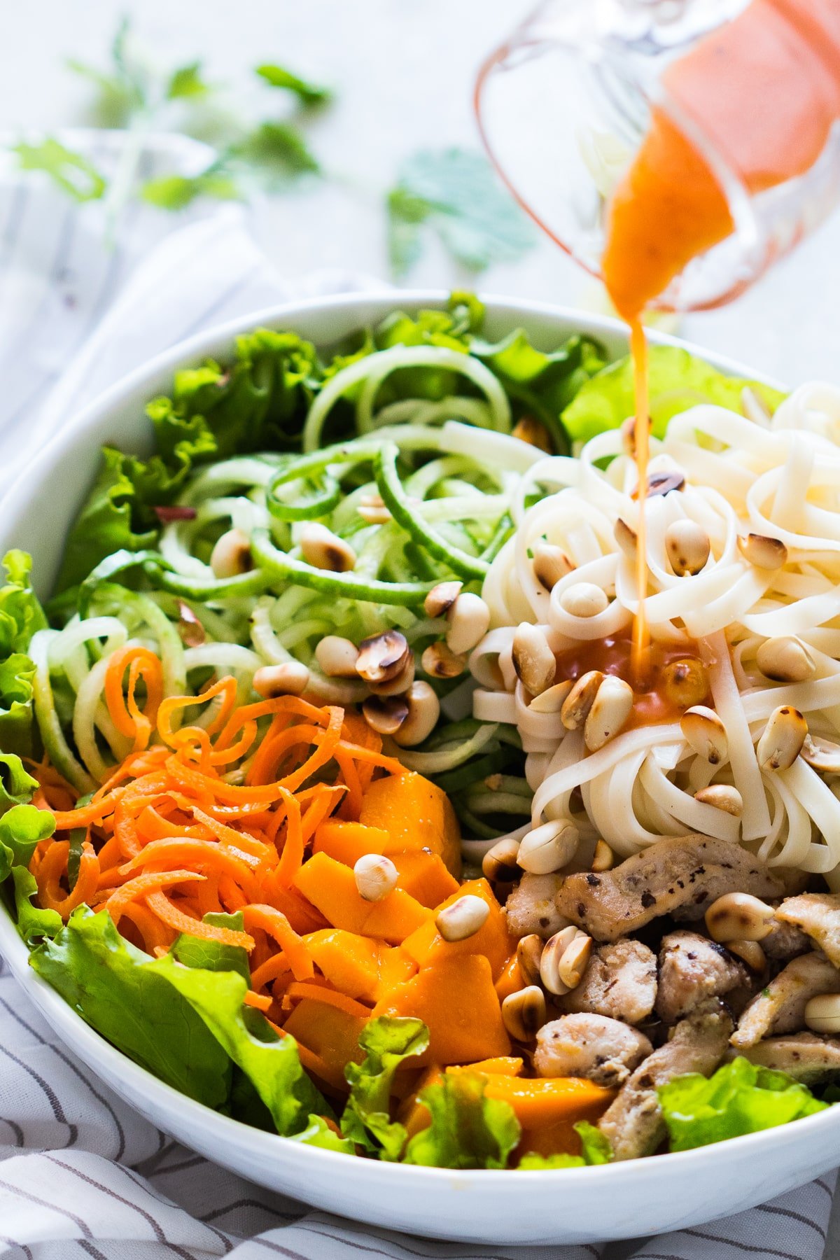 bowl of noodles, cucumber noodles, carrot noodles, mango, chicken, and lettuce and nuts with dressing poured over it