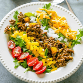 A plate with seasoned ground beef, shredded cheddar cheese, chopped onions, yellow bell peppers, cherry tomatoes, and leafy greens arranged in sections for a fresh low carb taco salad.