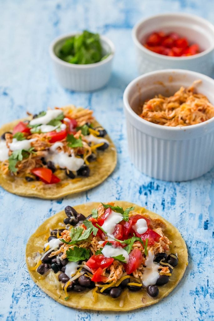 shredded chicken tacos with sour cream, cilantro, black beans, and salsa on them. Three bowls with cheese, cilantro, and tomato in them.