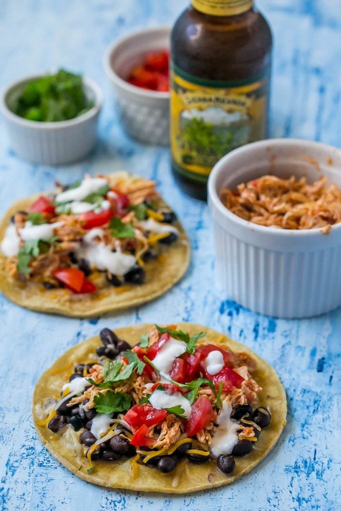 shredded chicken tacos with sour cream, cilantro, black beans, and salsa on them. Three bowls with cheese, cilantro, and tomato in them.