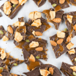 Close-up of Caramel Apple Chocolate Bark pieces topped with nuts, chunks of dried fruit, pumpkin seeds, and caramel swirls on parchment paper.