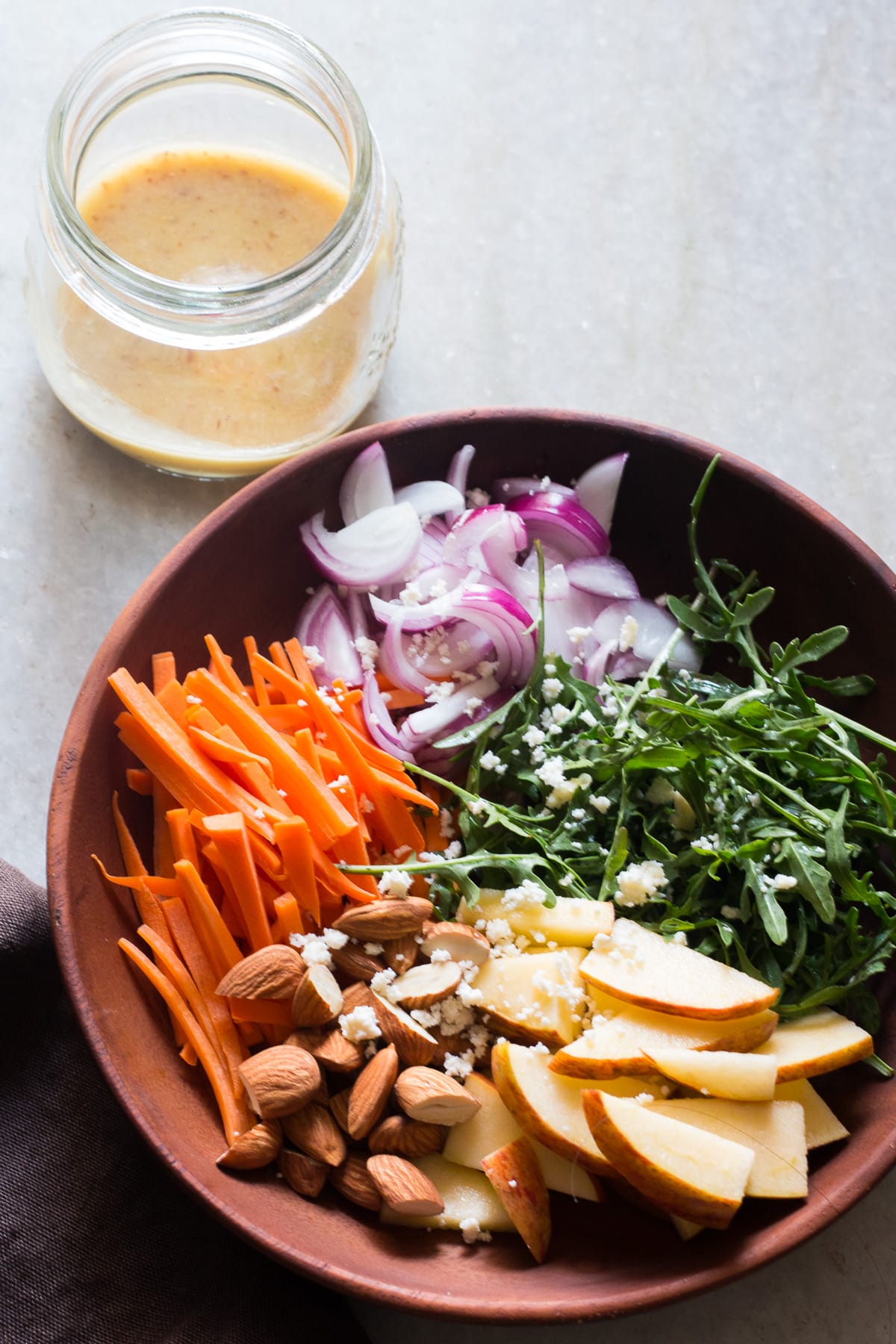 wooden bowl with apple slices, almonds, carrots, arugula, onion, and goat cheese in it