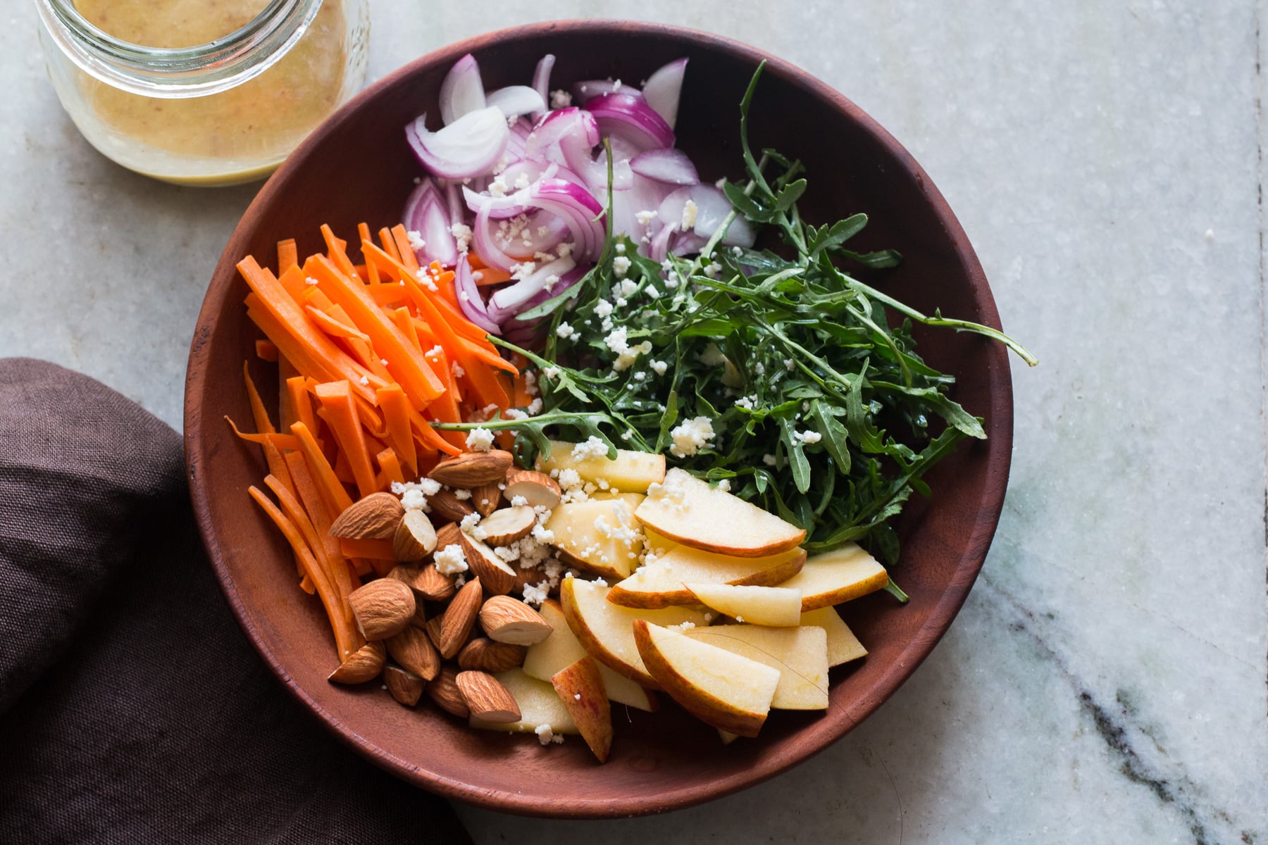 wooden bowl with apple slices, almonds, carrots, arugula, onion, and goat cheese in it