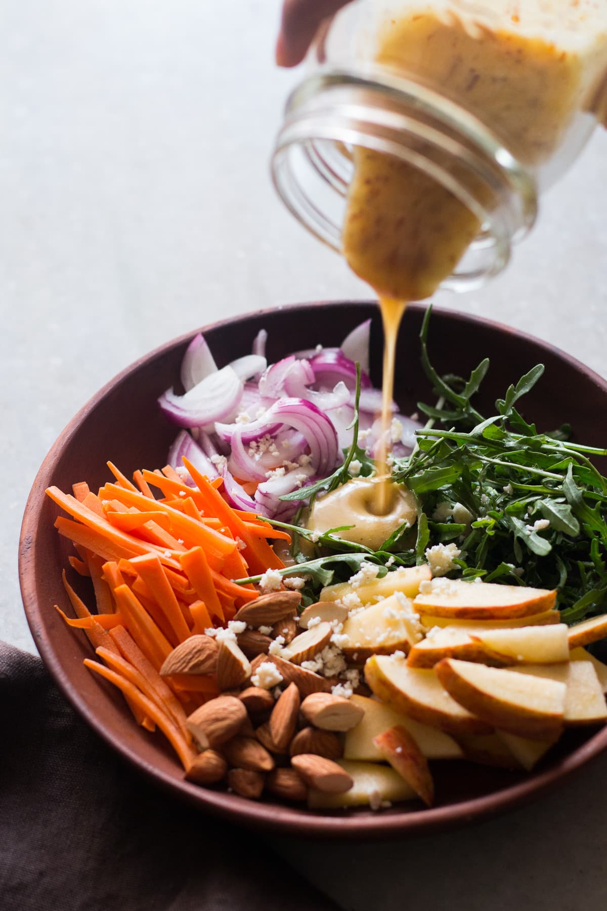 wooden bowl with apple slices, almonds, carrots, arugula, onion, and goat cheese in it, creamy dressing is being poured over it
