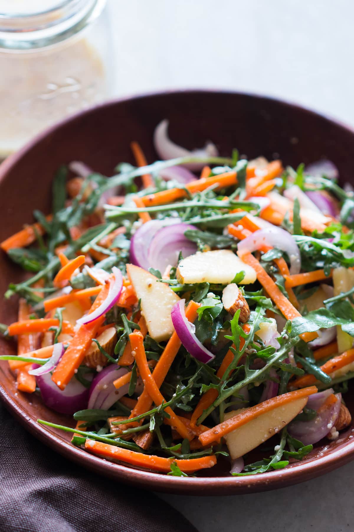 wooden bowl with apple slices, almonds, carrots, arugula, onion, and goat cheese in it all mixed up with creamy dressing