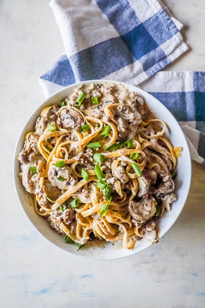 noodles with creamy stroganoff with mushrooms and chives on top sitting on a white plate blue plaid towel in background