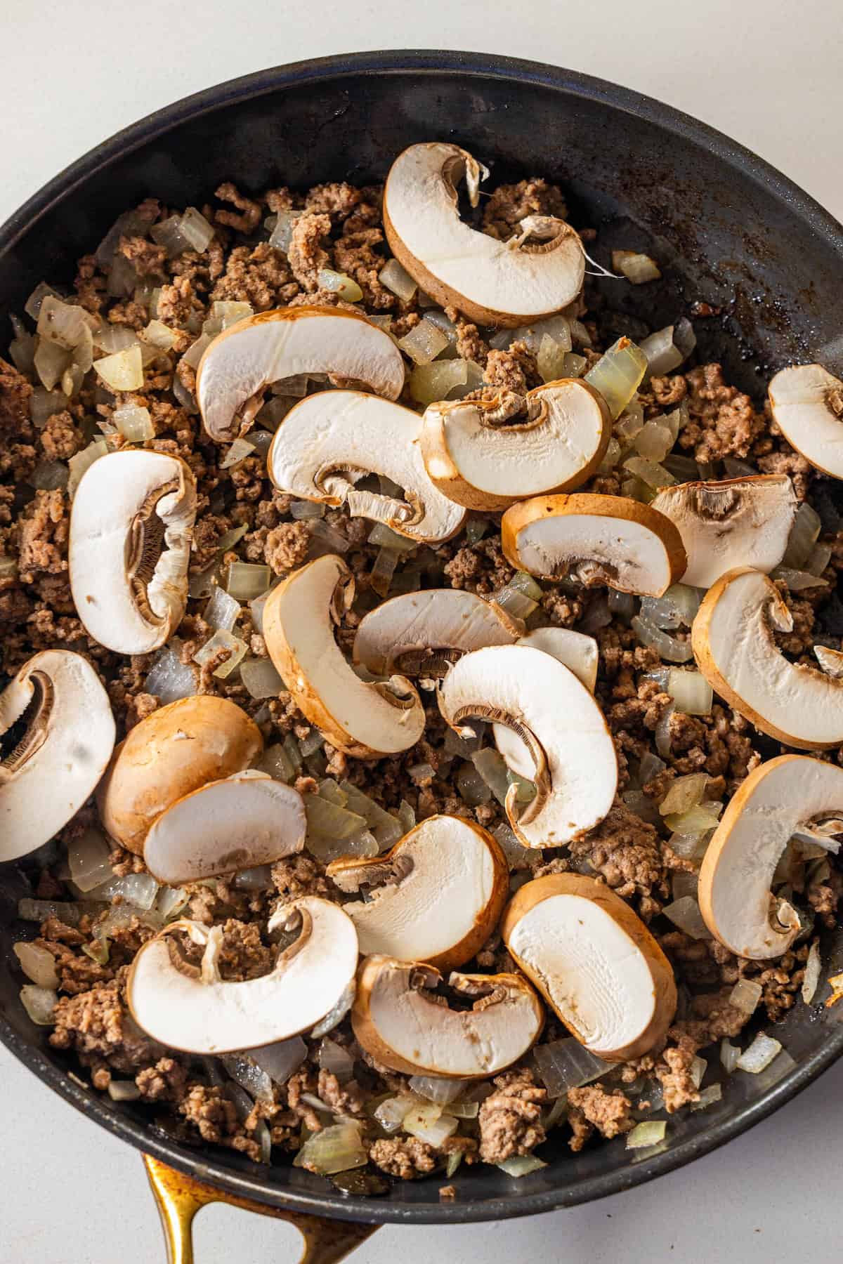 A skillet containing cooked ground beef and diced onions, topped with uncooked sliced mushrooms, perfect for starting a creamy Beef Stroganoff Fettuccine.