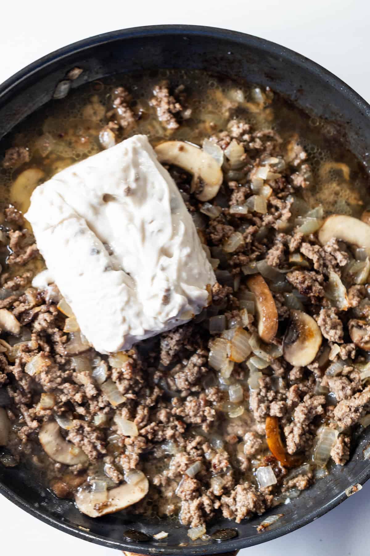 A skillet containing cooked ground beef, sliced mushrooms, diced onions, and a block of cream cheeseโthe perfect base for a Creamy Beef Stroganoff Fettuccine.