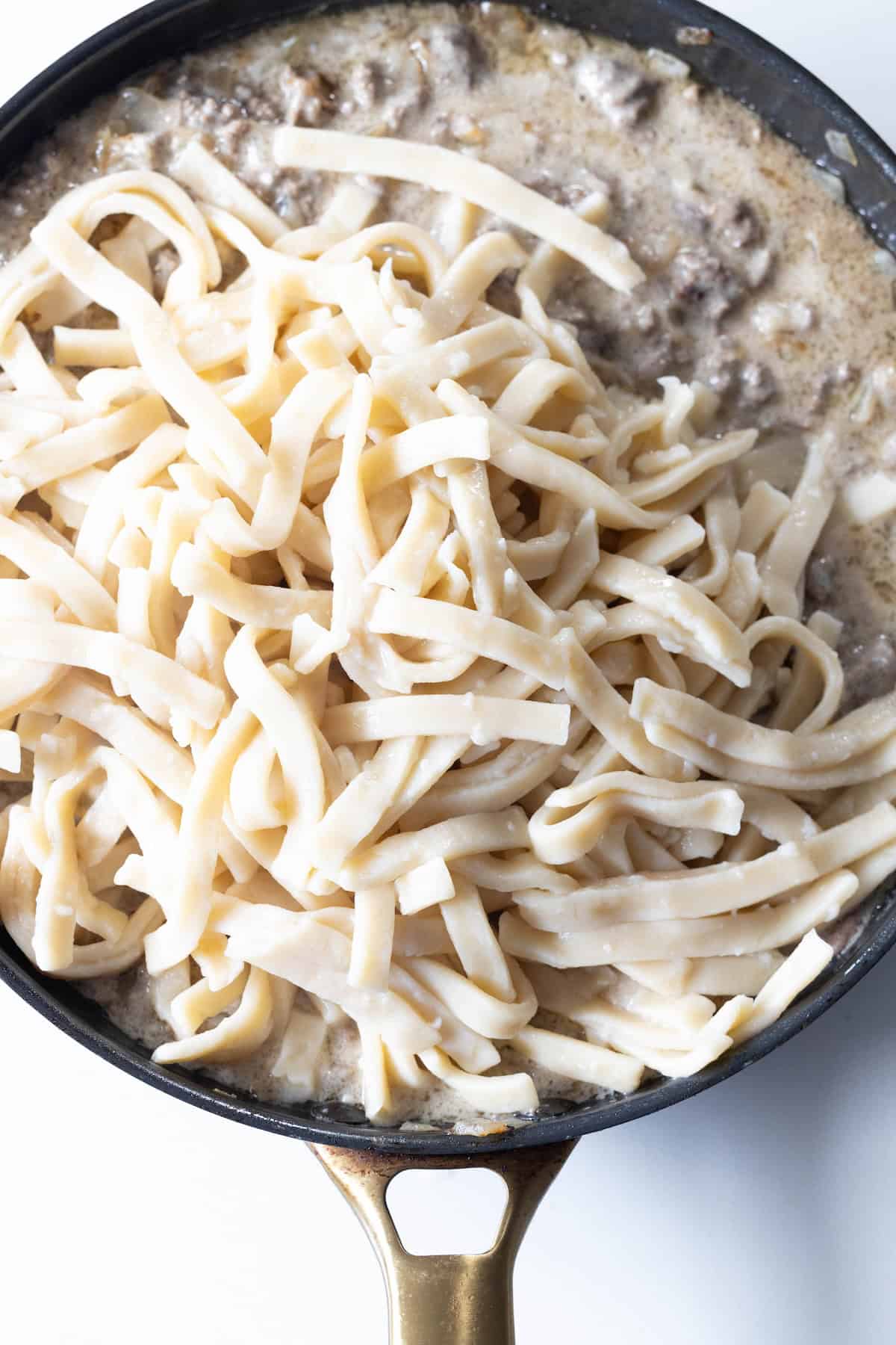 A skillet filled with creamy beef stroganoff fettuccine sauce and uncooked pasta, ready to simmer into a comforting meal.