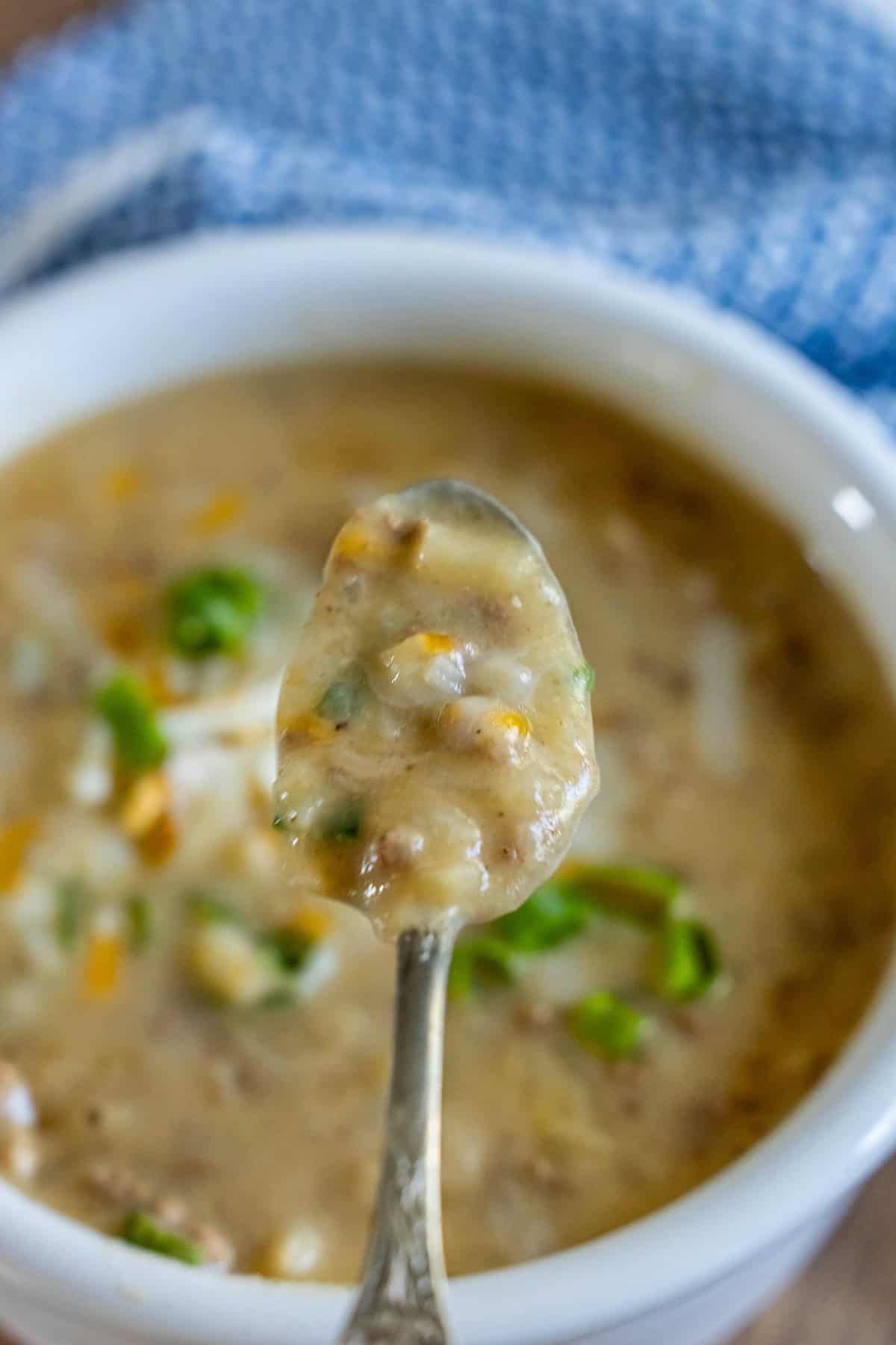 A spoonful of thick, creamy hash brown hamburger soup with visible herbs and vegetables is held over a white bowl on a wooden surface, with a blue cloth in the background.