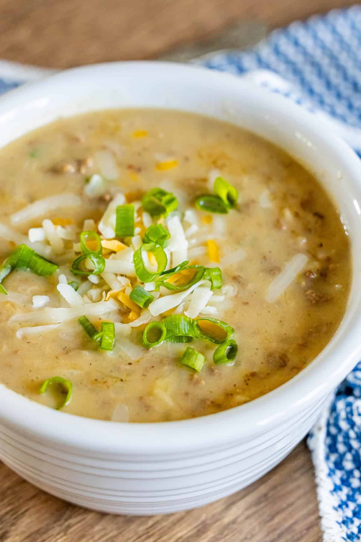 A bowl of creamy hash brown hamburger soup topped with shredded cheese and chopped green onions sits on a wooden table, next to a blue and white checkered cloth.