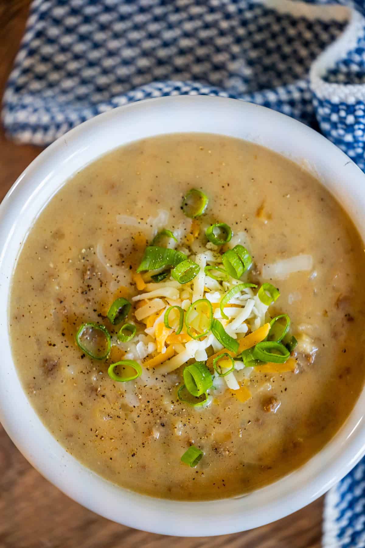 A bowl of creamy hash brown hamburger soup topped with shredded cheese and chopped green onions sits invitingly on a wooden surface, enhanced by the backdrop of a blue-patterned cloth.