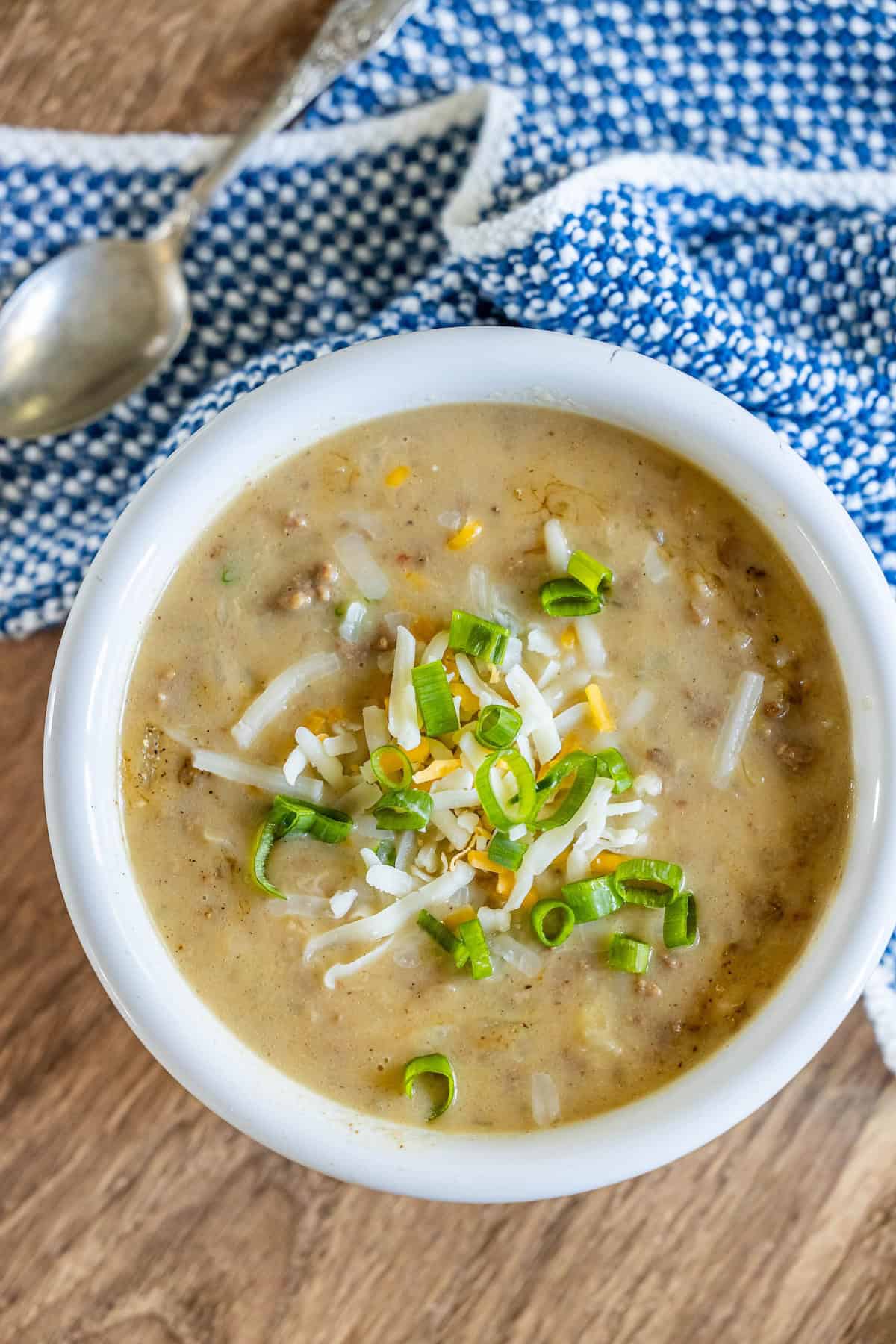 A bowl of creamy hash brown hamburger soup topped with shredded cheese and sliced green onions, placed on a wooden surface with a blue and white patterned cloth and a silver spoon nearby.