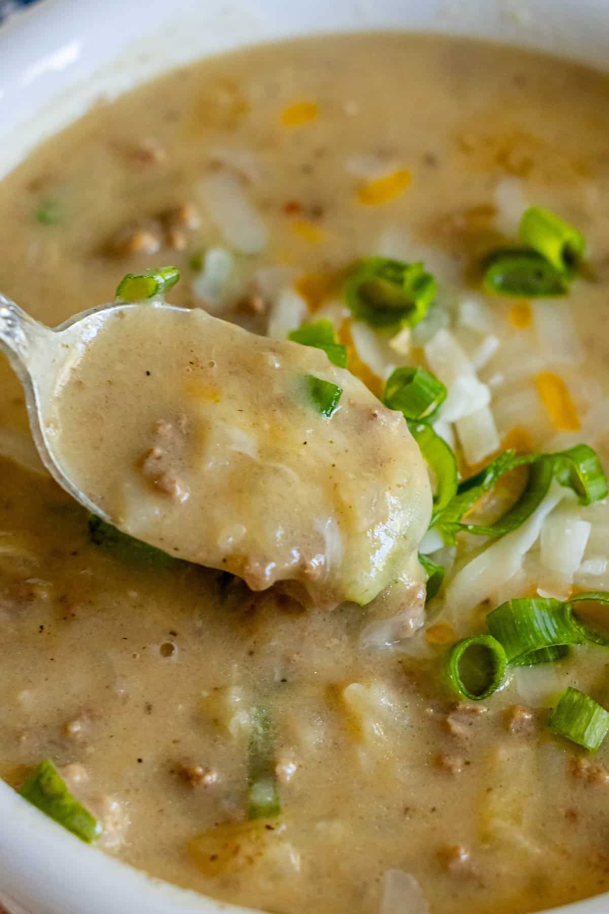 A close-up of creamy Hash Brown Hamburger Soup reveals chunky meat bits, topped with sliced green onions and shredded cheese. A spoon hovers above, poised with a luscious scoop of savory delight.