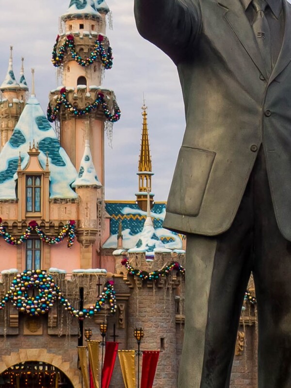 A festive statue of Walt Disney and Mickey Mouse adorned with Christmas decorations in front of a castle at Disneyland.