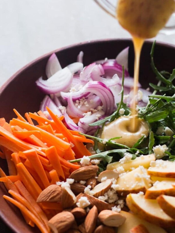 A bowl of salad with carrots, apples and a dressing being poured over it with arugula.