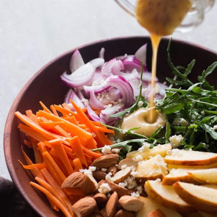A bowl of salad with carrots, apples and a dressing being poured over it with arugula.