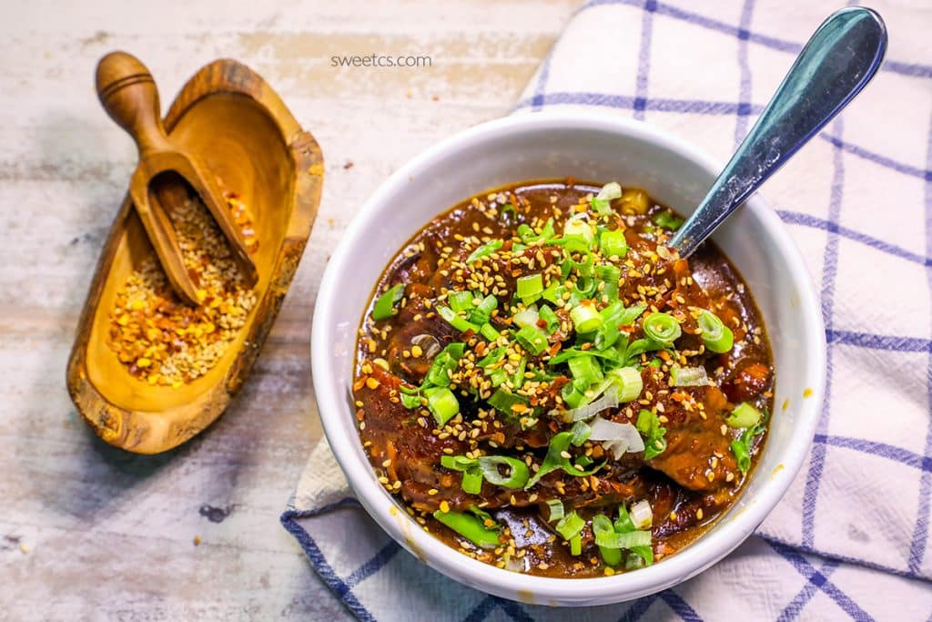 Mongolian beef stew in a bowl with green onions on it and a spoon in it with a wooden bowl and spoon and spices on the side. 
