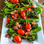 A white rectangular plate with sautéed broccolini, cherry tomatoes, and crispy pieces of bacon creates a vibrant bacon broccoli charred salad.