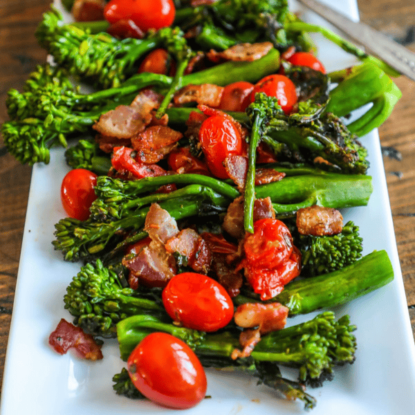 A white rectangular plate with sautéed broccolini, cherry tomatoes, and crispy pieces of bacon creates a vibrant bacon broccoli charred salad.