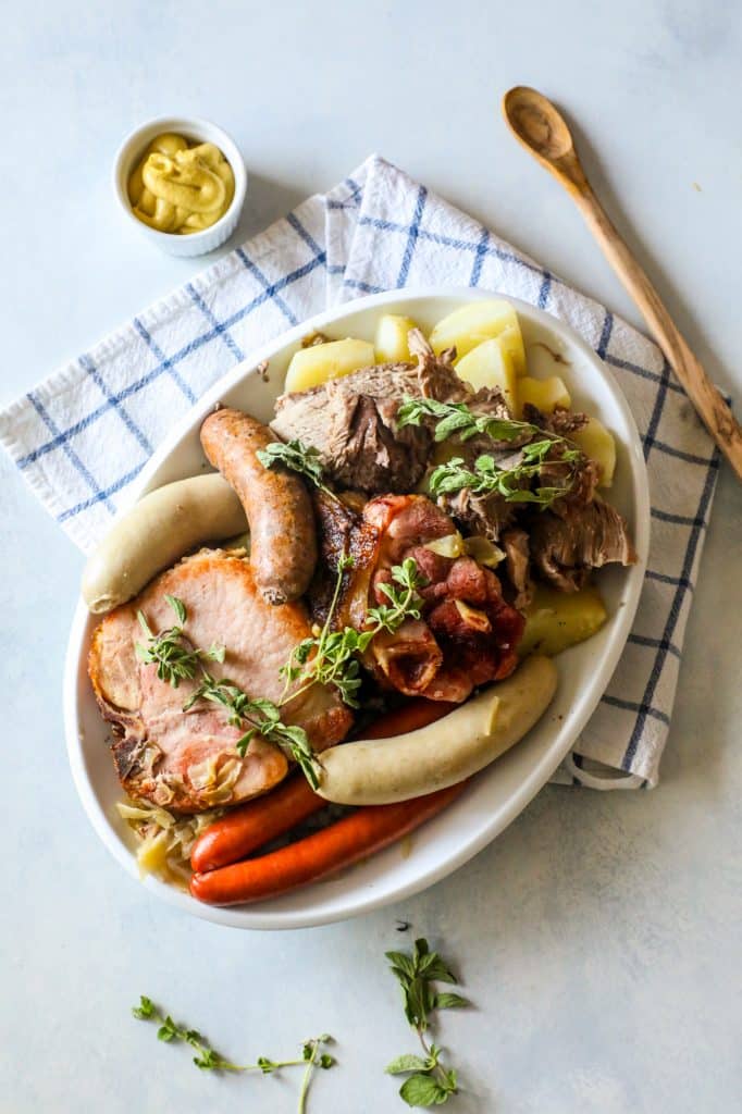 braised pork, ham, and sausages on a white plate with potatoes and thyme