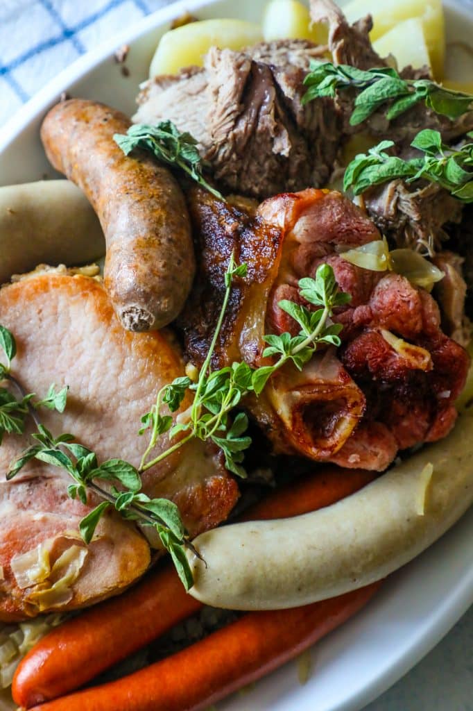 braised pork, ham, and sausages on a white plate with potatoes and thyme