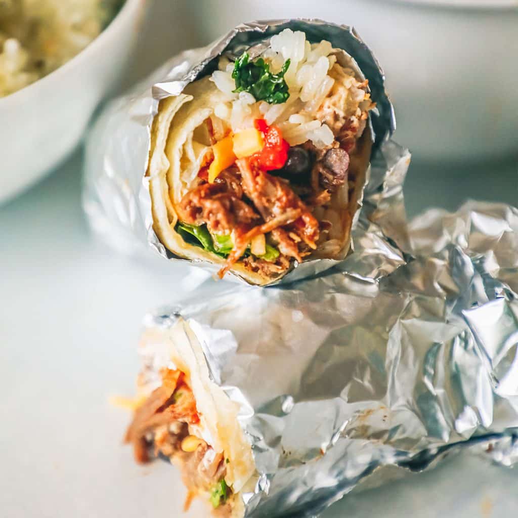 picture of barbacoa burritos wrapped in tin foil with a bowl of beans and rice in the background. 