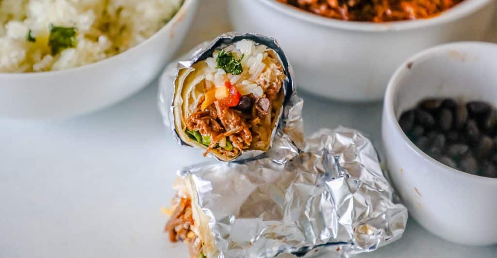 picture of barbacoa burritos wrapped in tin foil with a bowl of beans and rice in the background. 