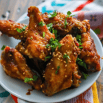 A plate of Sweet and Spicy Crunchy Baked Wings, glazed and garnished with chopped herbs and sesame seeds, sits on a table with a colorful cloth in the background.