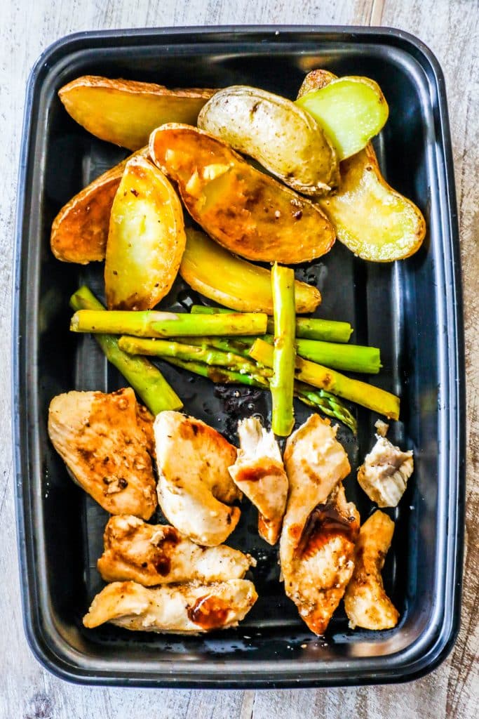plastic container with roasted potatoes, asparagus, and cooked chicken in it