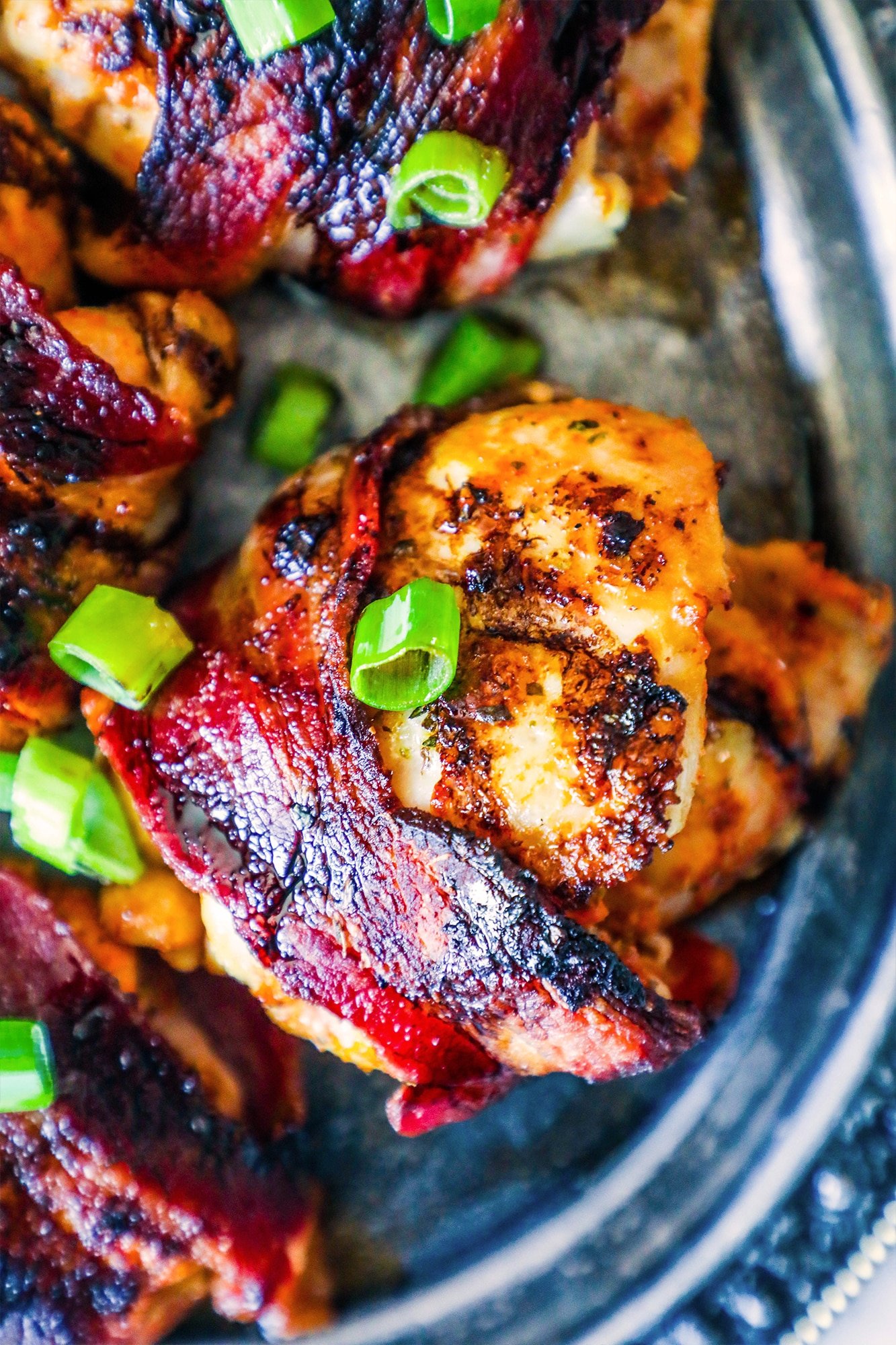 bacon wrapped chicken thighs on a silver plate with green onions on them up close. 