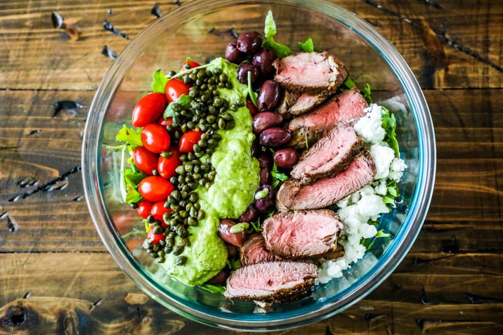 bowl of steak, capers, tomatoes, olives, cheese, and lettuce with green dressing