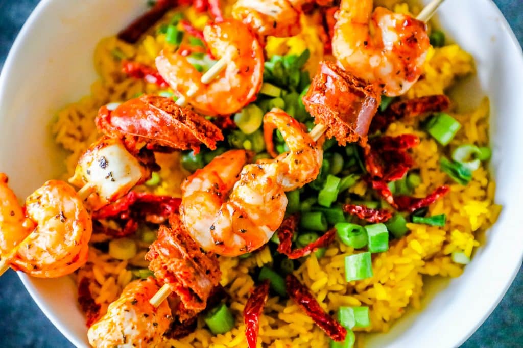 shrimp, chicken, and sausage on a skewer over yellow rice with sundried tomatoes and green onions