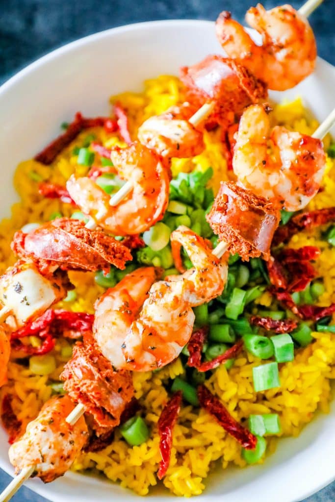 shrimp, chicken, and sausage on a skewer over yellow rice with sundried tomatoes and green onions
