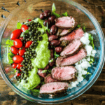 A bowl filled with sliced beef, cherry tomatoes, kalamata olives, lentils, cauliflower rice, and a dollop of creamy avocado sauce.