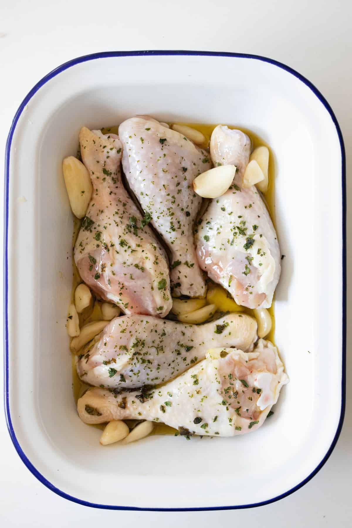 Greek chicken drumsticks marinated with herbs and garlic cloves in a white baking dish.