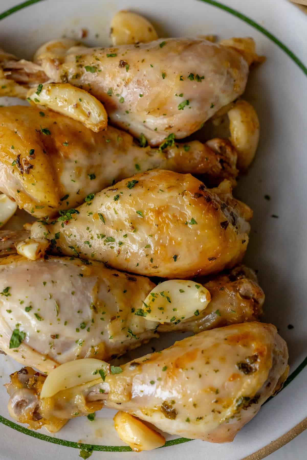 Greek chicken drumsticks, cooked to perfection, are garnished with chopped herbs and fragrant garlic cloves on a plate.